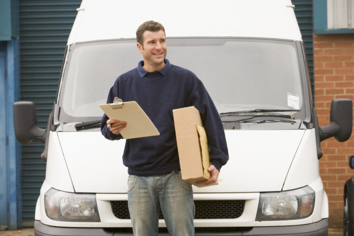 Deliveryperson standing with van holding clipboard and box smili
